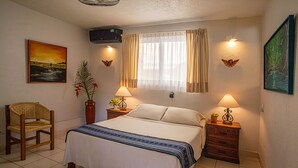 Un dormitorio con una cama, dos mesitas de noche con lámparas, una silla, un ventanal con cortinas y una obra de arte en la pared.