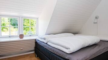 A bedroom with a sloped ceiling, a window with a view of greenery, a bed with white bedding, and a wall-mounted light fixture.