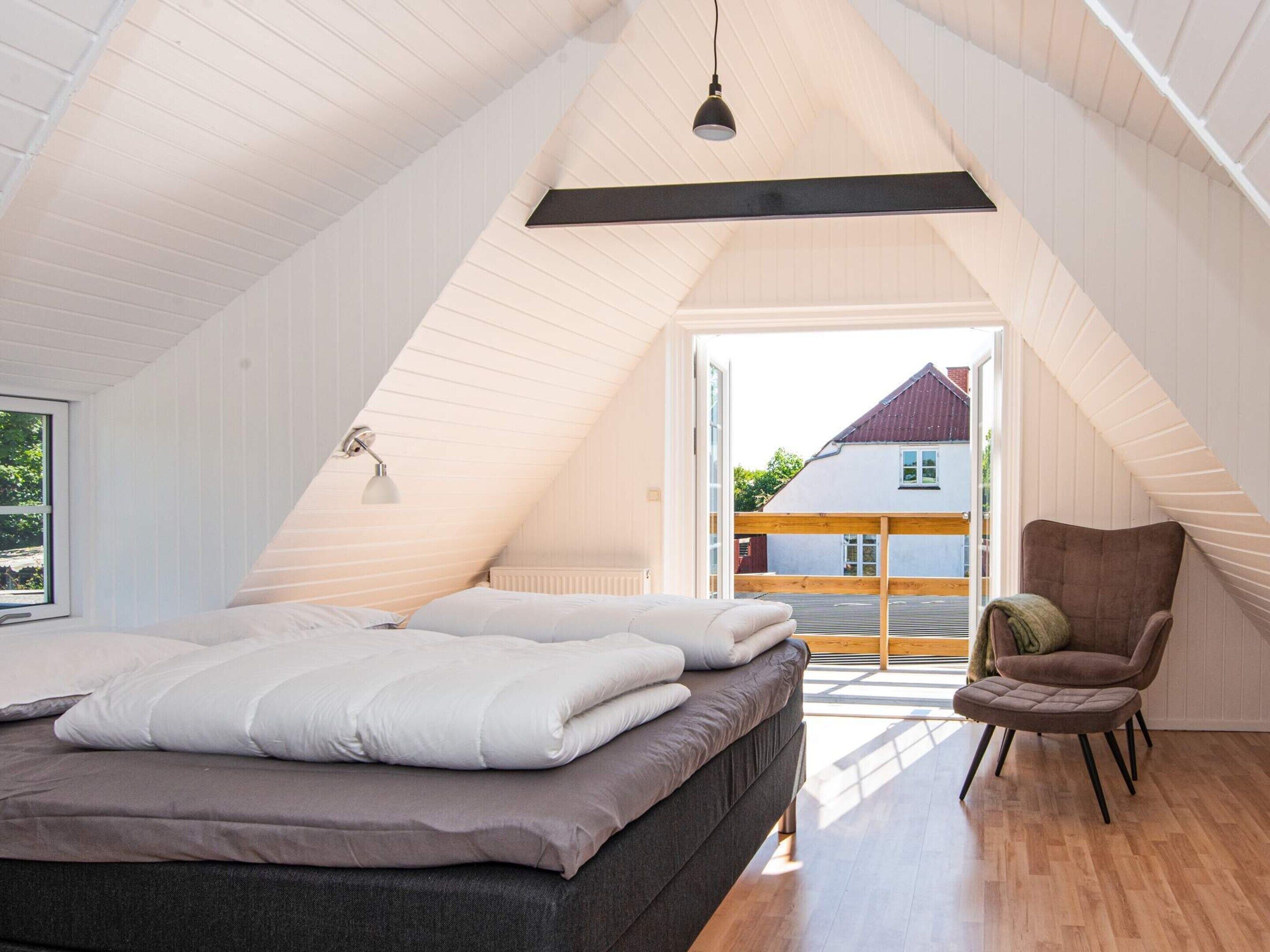 Un dormitorio con techo inclinado, una cama con almohadas, una silla y vistas al exterior.