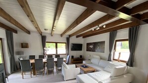 Un salon spacieux avec un plafond en bois, un canapé blanc, une table à manger avec des chaises, un téléviseur et de grandes fenêtres.
