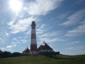 Um farol com torre listrada em vermelho e branco, um edifício branco com telhado vermelho e uma colina gramada em primeiro plano.