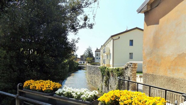 A canal with flower planters on a bridge, buildings in the background, and a stone wall on one side. - Apartment in Garda Near Lake (Garda)