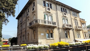 A multi-story building with balconies, large windows, and a street in front with flower planters. - Apartment in Garda Near Lake (Garda)