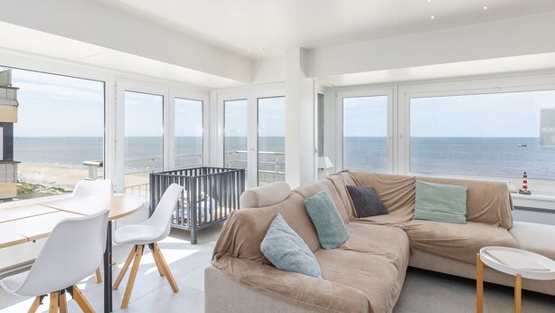 A spacious living area with a large window overlooking the sea, a beige sofa with cushions, a white table, and a white chair. - Bright Apartment With Frontal sea View (Middelkerke)