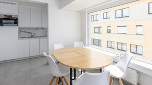 A modern kitchen with white cabinets, a marble countertop, and an oven. There is a dining area with a wooden table and white chairs. - Bright Apartment With Frontal sea View (Middelkerke)