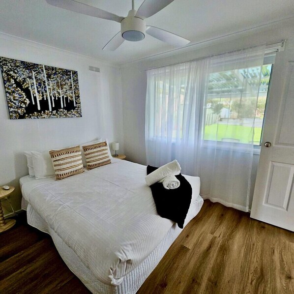 A bedroom with a large bed, a ceiling fan, a painting on the wall, and a window with sheer curtains. - Prom Coast - Duneside (Sandy Point)