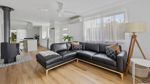 A modern living room with a black leather sectional sofa, a small white table, and a floor lamp. There is a kitchen area in the background with white cabinets and a bar stool.