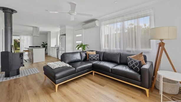 A modern living room with a black leather sectional sofa, a small white table, and a floor lamp. There is a kitchen area in the background with white cabinets and a bar stool. - Prom Coast - Duneside (Sandy Point)