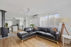 Una sala de estar moderna con un sofá seccional de cuero negro, piso de madera, ventilador de techo y un gran ventanal con cortinas translúcidas.