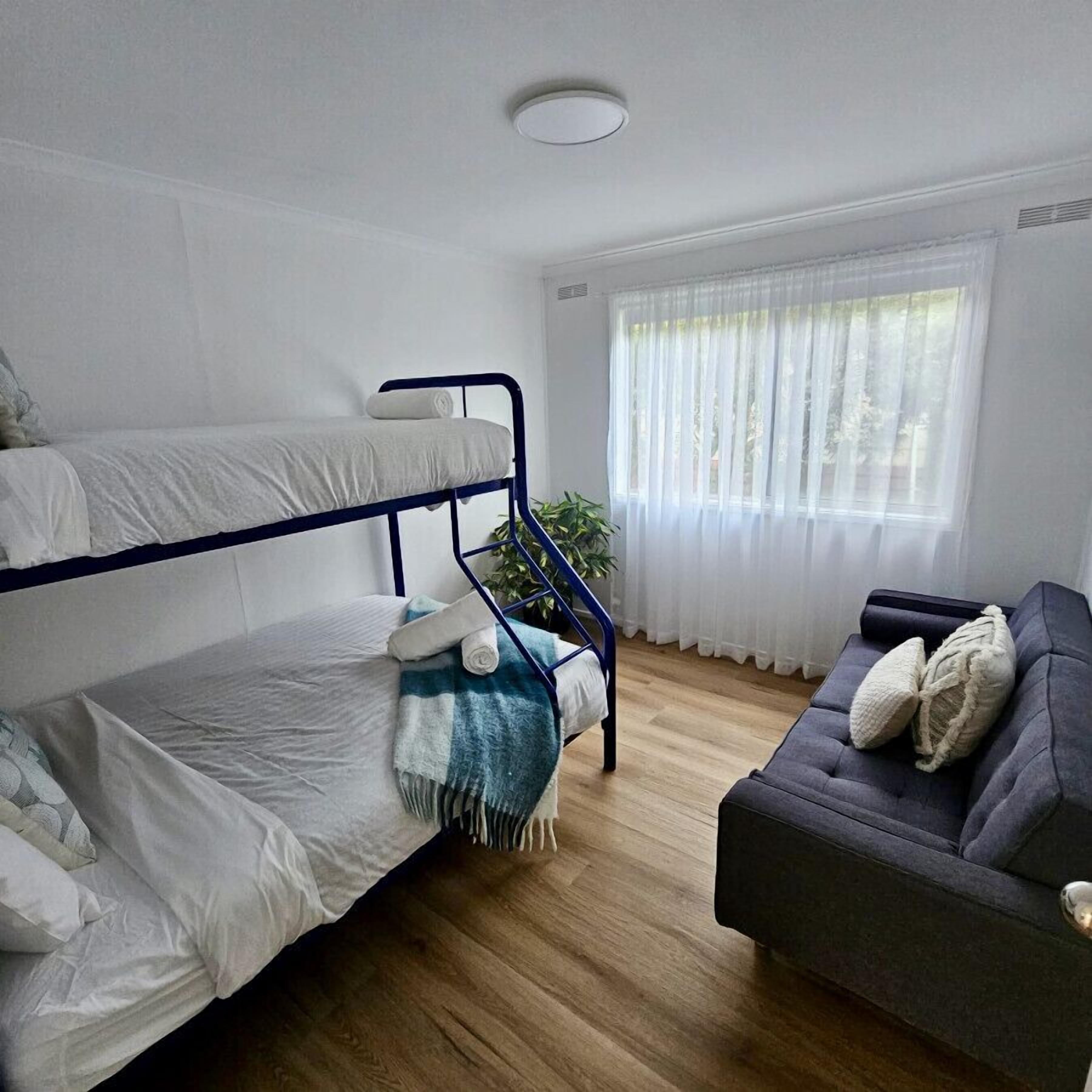A bunk bed with a blue frame, a sofa, a window with sheer curtains, and a ceiling light.
