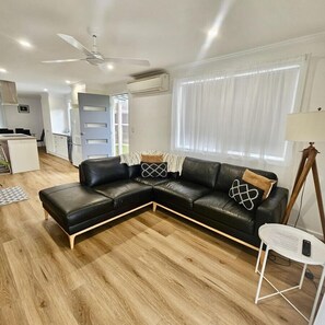 A modern living room with a black leather sectional sofa, a dining area with a white table and chairs, and a kitchen in the background. - Prom Coast - Duneside (Sandy Point)