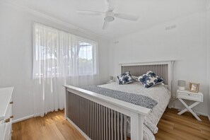 A bedroom with a bed, a bedside table, a nightstand, a ceiling fan, and a window with sheer curtains. - Prom Coast - Duneside (Sandy Point)