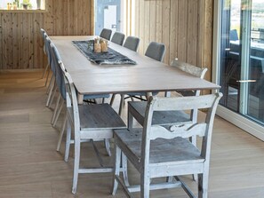 Une grande table à manger en bois, entourée de six chaises, se trouve dans une pièce aux murs en bois et aux grandes fenêtres.