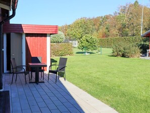 A patio with a table and chairs, a red building, and a well-maintained lawn with trees and a hedge.