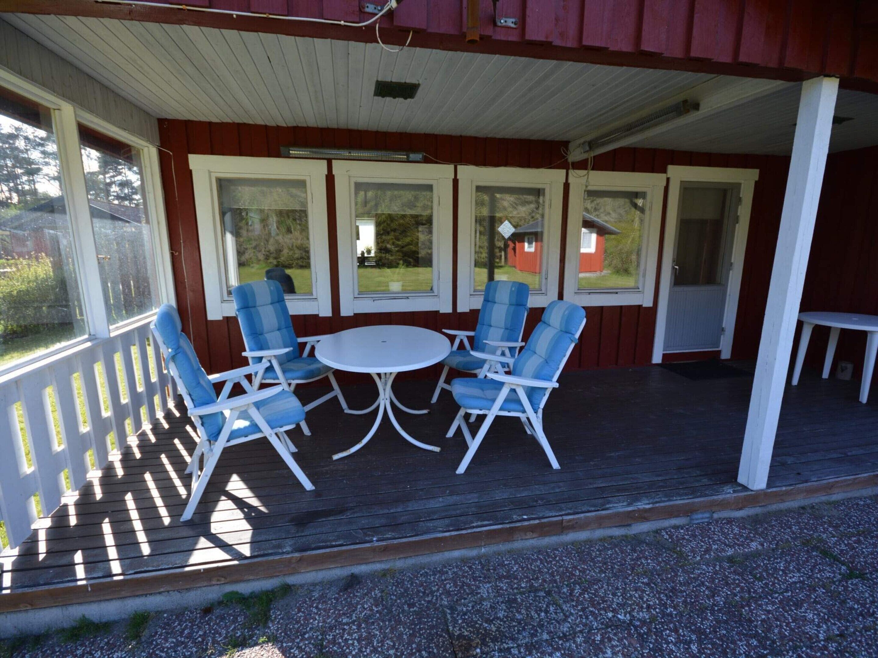 A covered outdoor seating area with a round table and four chairs, a wooden deck, and a view of a red building through large windows.