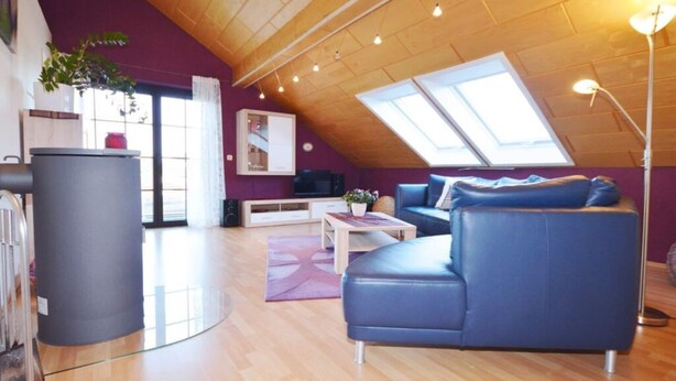 A modern loft-style living room with a sloped ceiling, a blue sofa, a coffee table, and a large window providing natural light. - Holiday Apartment ML Lafos (Konz)