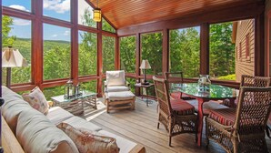 A spacious deck with wooden flooring, wicker furniture, and large windows offering a view of a forested landscape. - Schoolmarm Rock - 450' of private waterfront on Rangeley Lake (Rangeley)