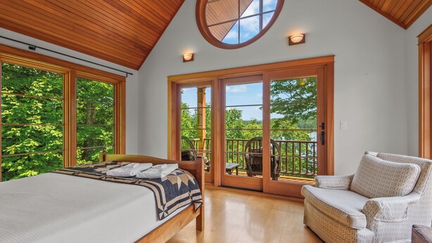 A bedroom with a large bed, a chair, and a balcony with a view of trees. - Schoolmarm Rock - 450' of private waterfront on Rangeley Lake (Rangeley)