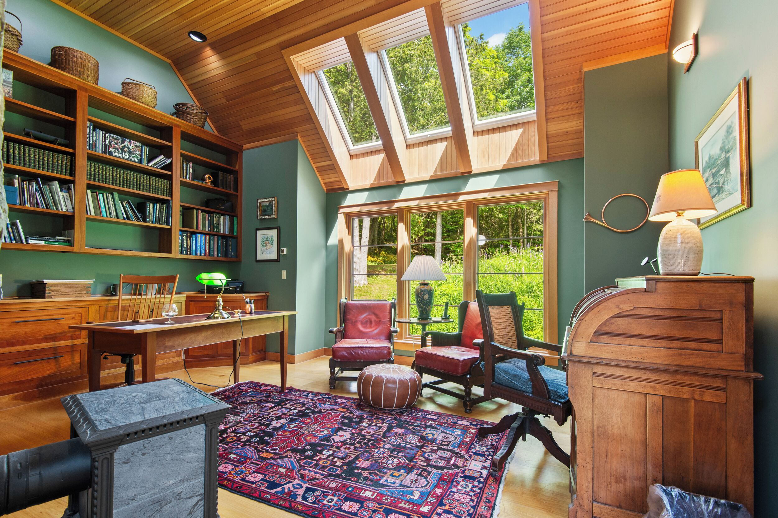 Un salón de lectura acogedor con techo de madera, tragaluces, una amplia estantería, asientos cómodos y vistas al exterior.