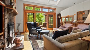 A spacious living room with a fireplace, leather chairs, a sofa, and a large window with a view of greenery. - Schoolmarm Rock - 450' of private waterfront on Rangeley Lake (Rangeley)
