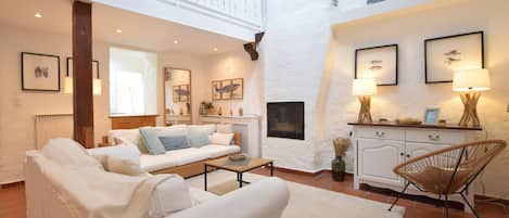 A spacious living area with a high ceiling, white walls, and a fireplace. There are two sofas, a coffee table, and a wicker chair. The room has framed pictures on the walls and a lamp on a side table.