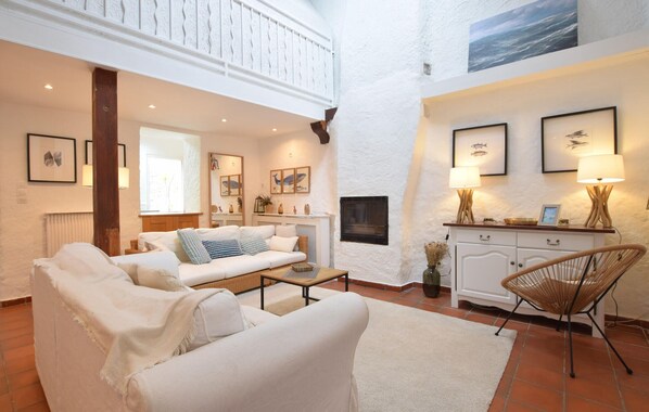 Amplia sala de estar con techo alto, paredes blancas y chimenea. Hay dos sofás, una mesa de centro y un sillón de mimbre. La habitación tiene cuadros en las paredes y una lámpara sobre una mesita auxiliar.