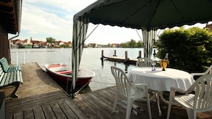 A wooden deck with a table and chairs, a small boat, and a view of a river with buildings in the background.