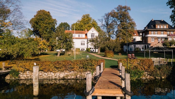 A wooden dock leading to a house with a garden and a pond.