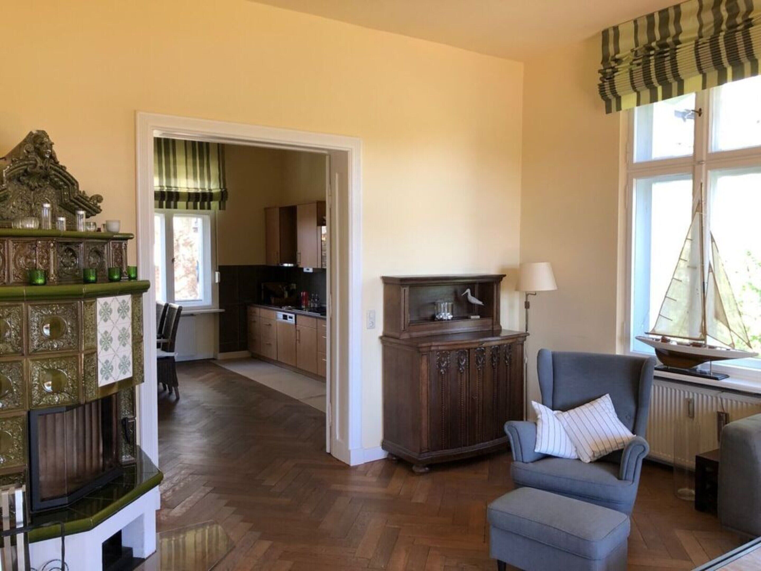 A spacious living area with a wooden floor, a blue armchair, a decorative cabinet, and a view of a kitchen through an open doorway.