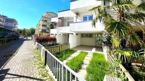 Un edifici residencial amb diversos balcons, un camí pavimentat i una petita zona de jardí amb gespa i plantes.