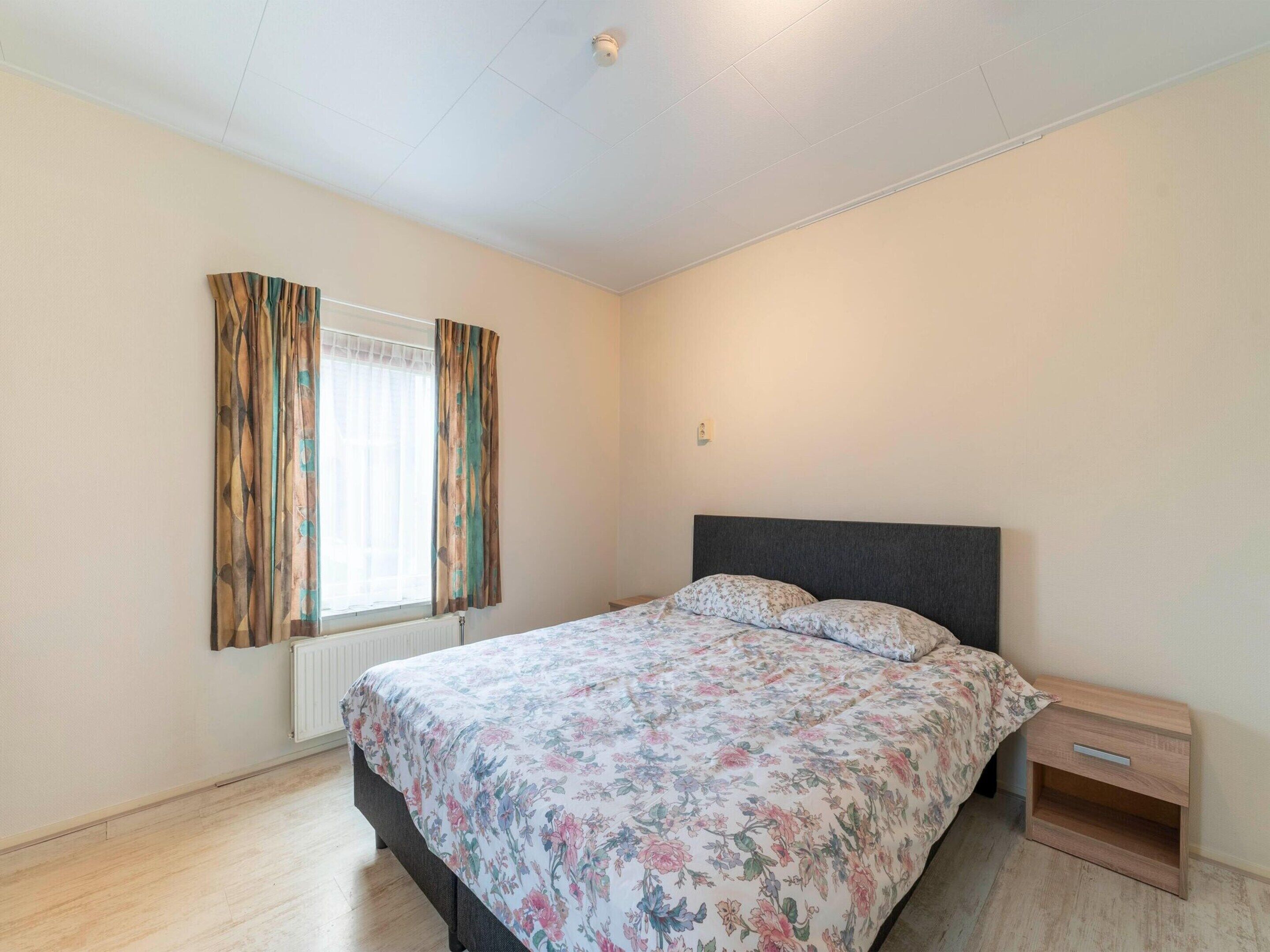 A bedroom with a bed, a nightstand, a window with curtains, and a radiator.