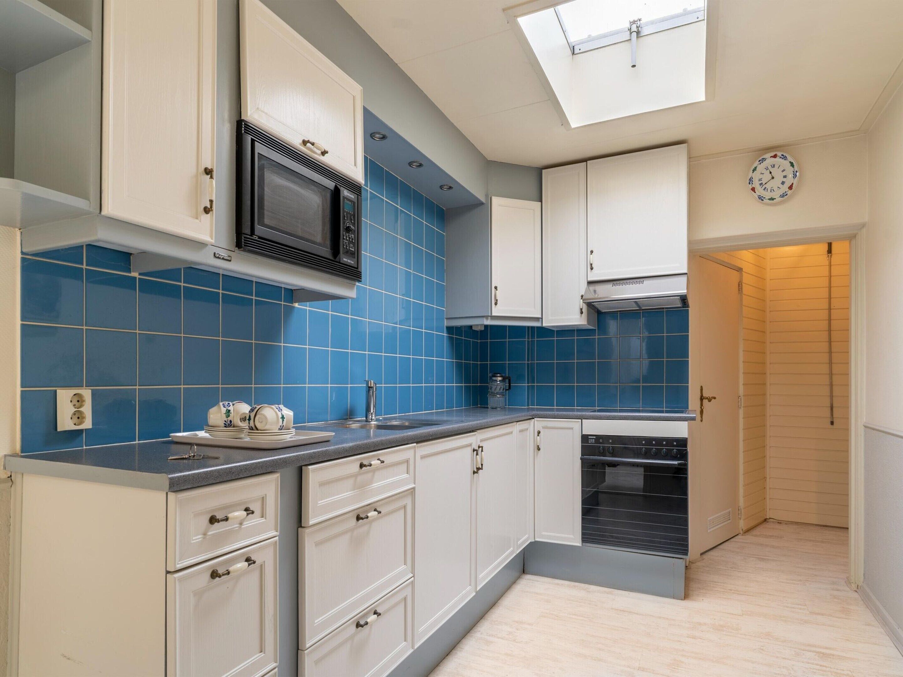 Una cocina con armarios blancos, azulejos azules, un microondas, un lavavajillas y un tragaluz.