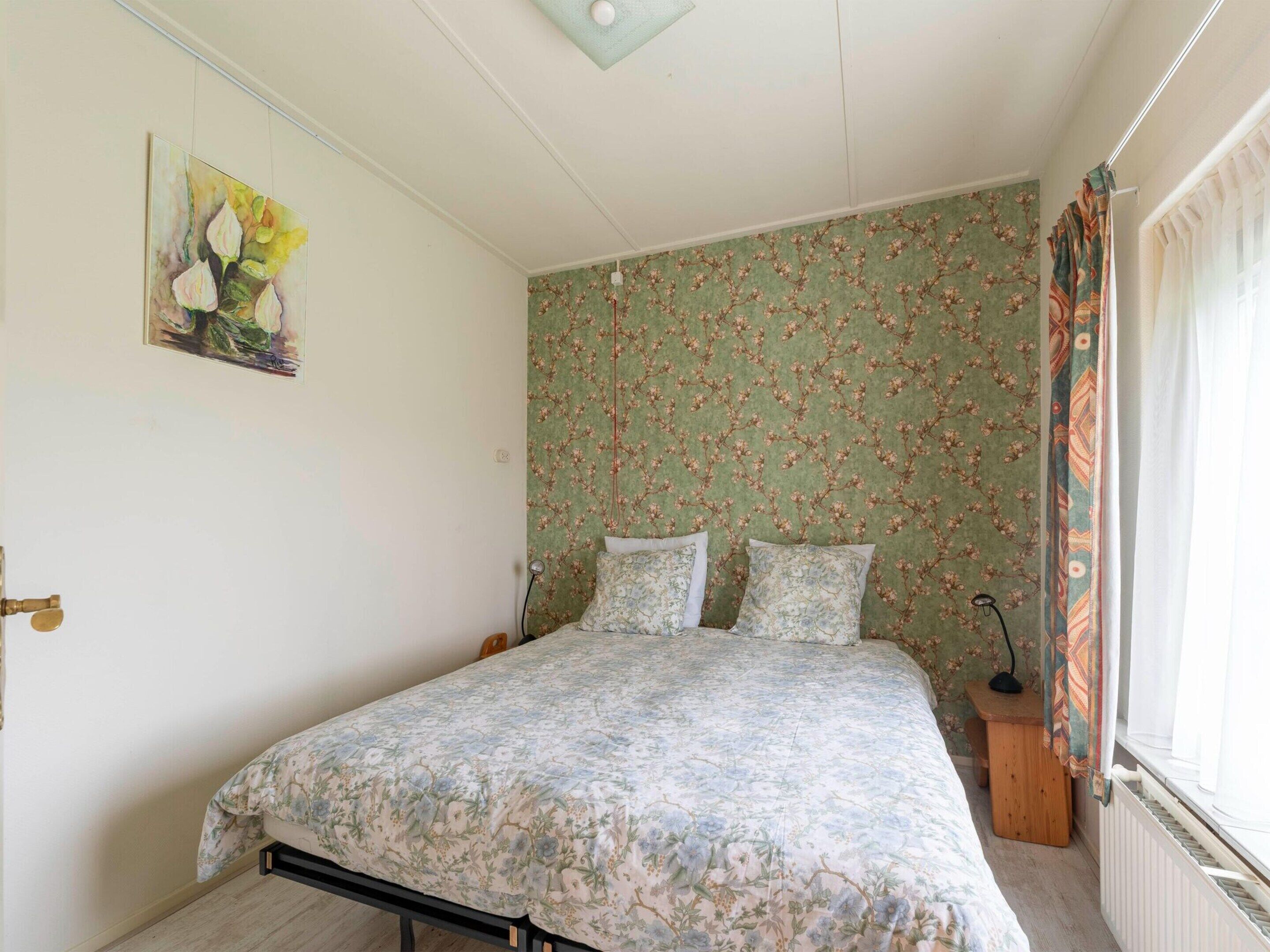 A bedroom with a floral bedspread, a painting on the wall, and a window with curtains.