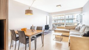 A modern living room with a dining area, a sofa, a TV, and a large window offering a view of the city. - Family Stay in Oostduinkerke (Koksijde)
