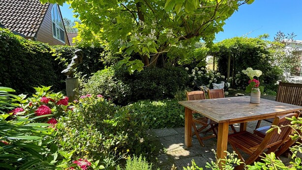 A wooden table and chairs set outdoors in a garden with various plants and flowers.