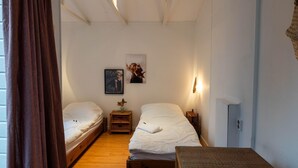 A room with two single beds, a small wooden bench, a wall-mounted shelf with a vase, and a ceiling light fixture.