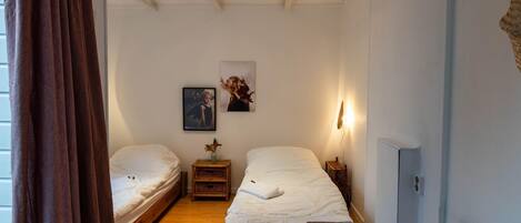 Une chambre avec deux lits simples, un petit banc en bois, une étagère fixée au mur avec un vase, et un luminaire au plafond.