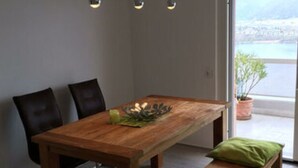A modern dining area with a wooden table, two chairs, and a bench. There are pendant lights above the table and a view of a body of water through a large window. - Holiday Apartment Casa Chimp 6 (Gambarogno)