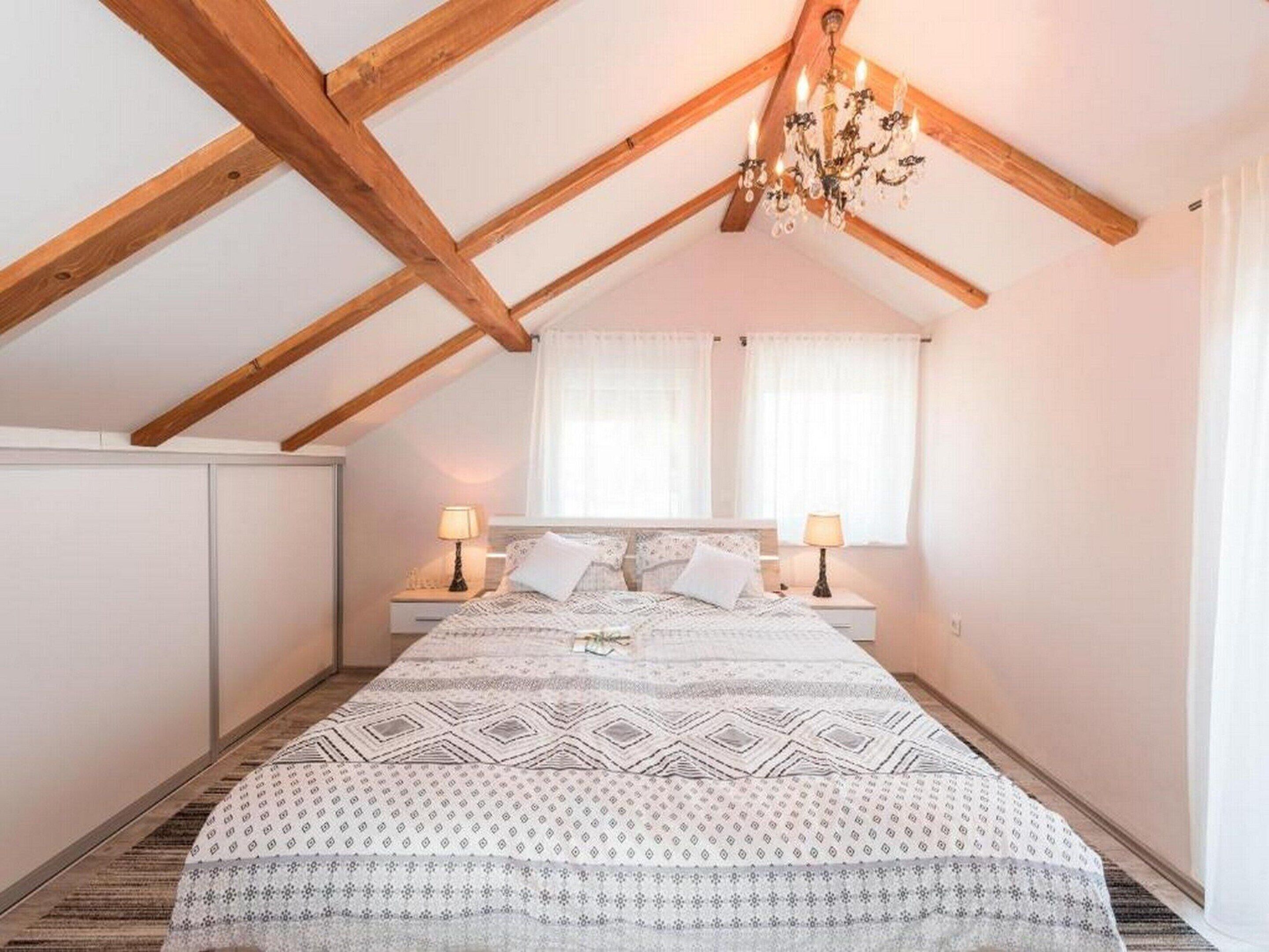 Ein Schlafzimmer mit schrägen Wänden, Holzbalken, einem Bett mit gemusterten Textilien, zwei Nachttischlampen und einer Kristallleuchter.