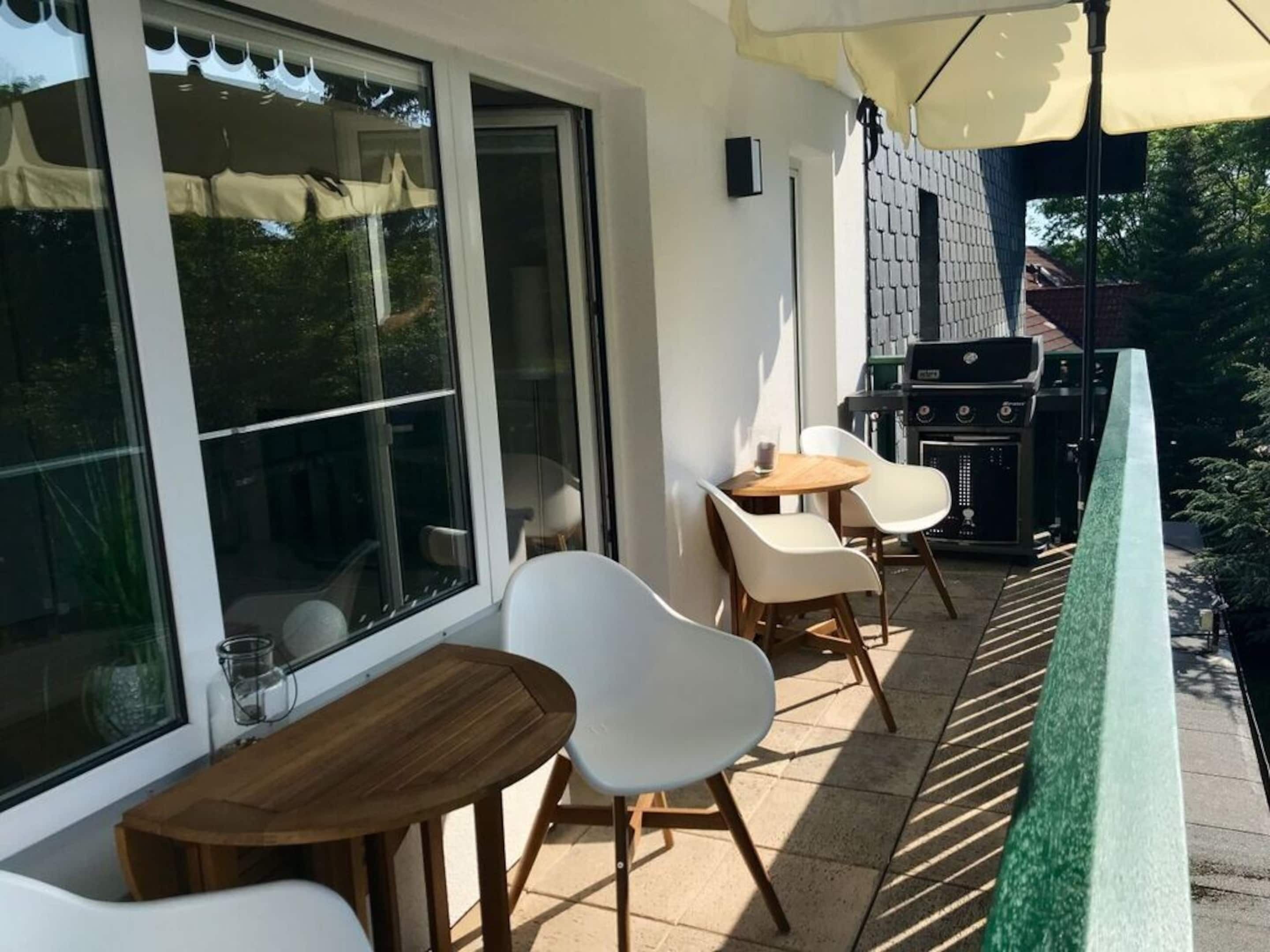 A patio area with a wooden table and white chairs, a grill, and a glass door leading to a garden.