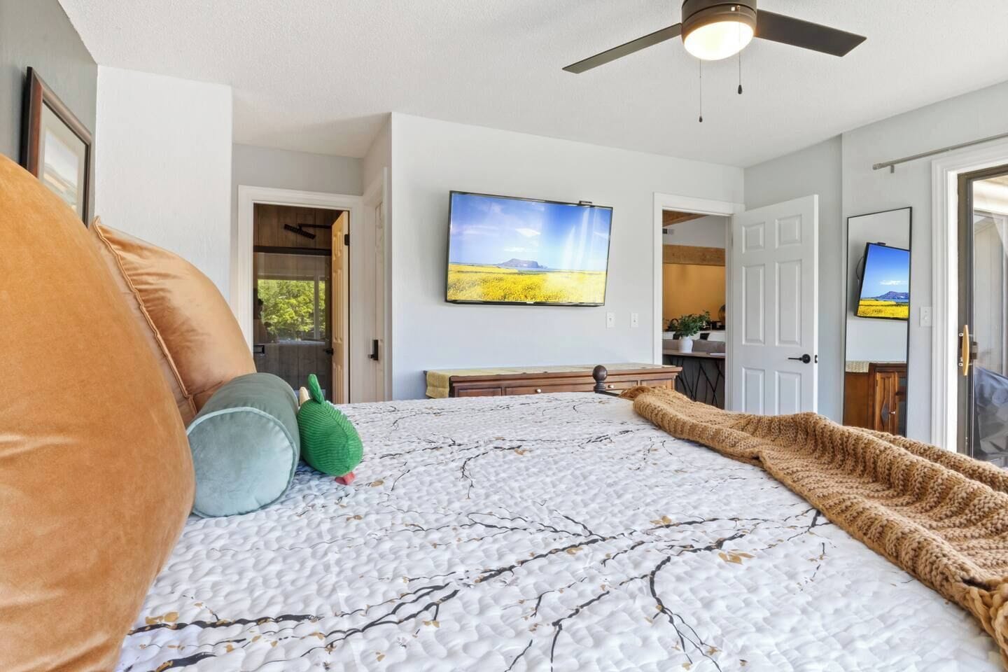 Ein Schlafzimmer mit Bett, Kissen, Decke, Deckenventilator, Fernseher und einem Gemälde einer Landschaft.