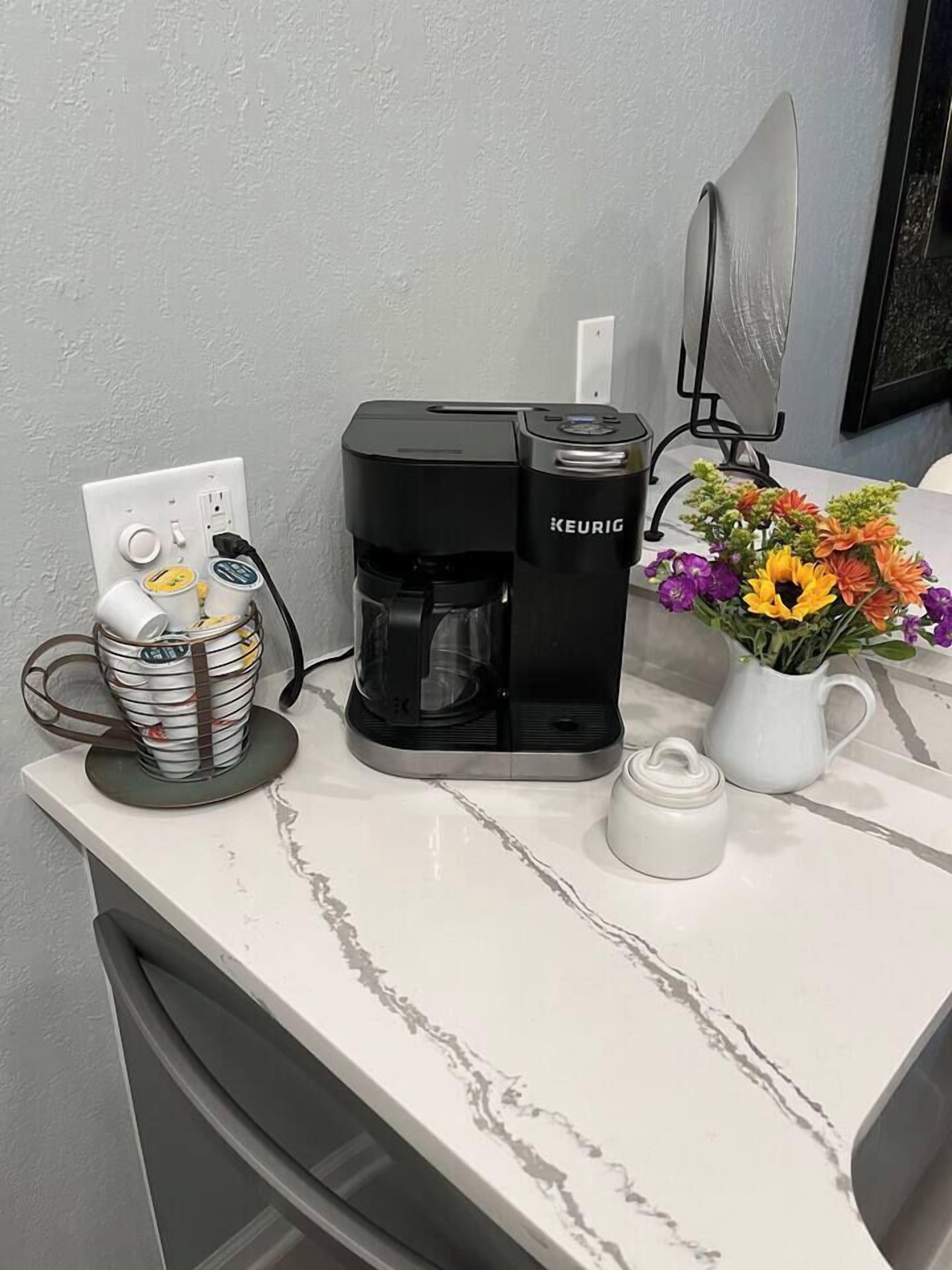 En kaffebryggare står på en vit bänkskiva bredvid ett eluttag. Bredvid finns en korg med koppar och en vas med blommor.