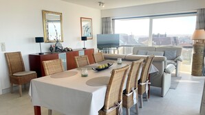 Un espace de repas moderne, avec une table dressée pour six personnes, entourée de chaises en osier. Un canapé et un téléviseur sont visibles au fond, et de grandes fenêtres offrent une vue sur la ville.