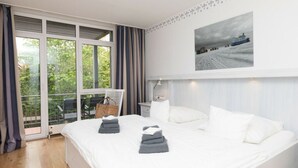 Ein Schlafzimmer mit einem großen Bett, einem Fenster mit Vorhängen, einem Sessel, einem Tisch und einem Bild an der Wand.