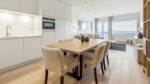 A modern kitchen with built-in appliances, a dining area with a large table and chairs, and a view of the sea through large windows. - Apartment Next to the Casino (Blankenberge)