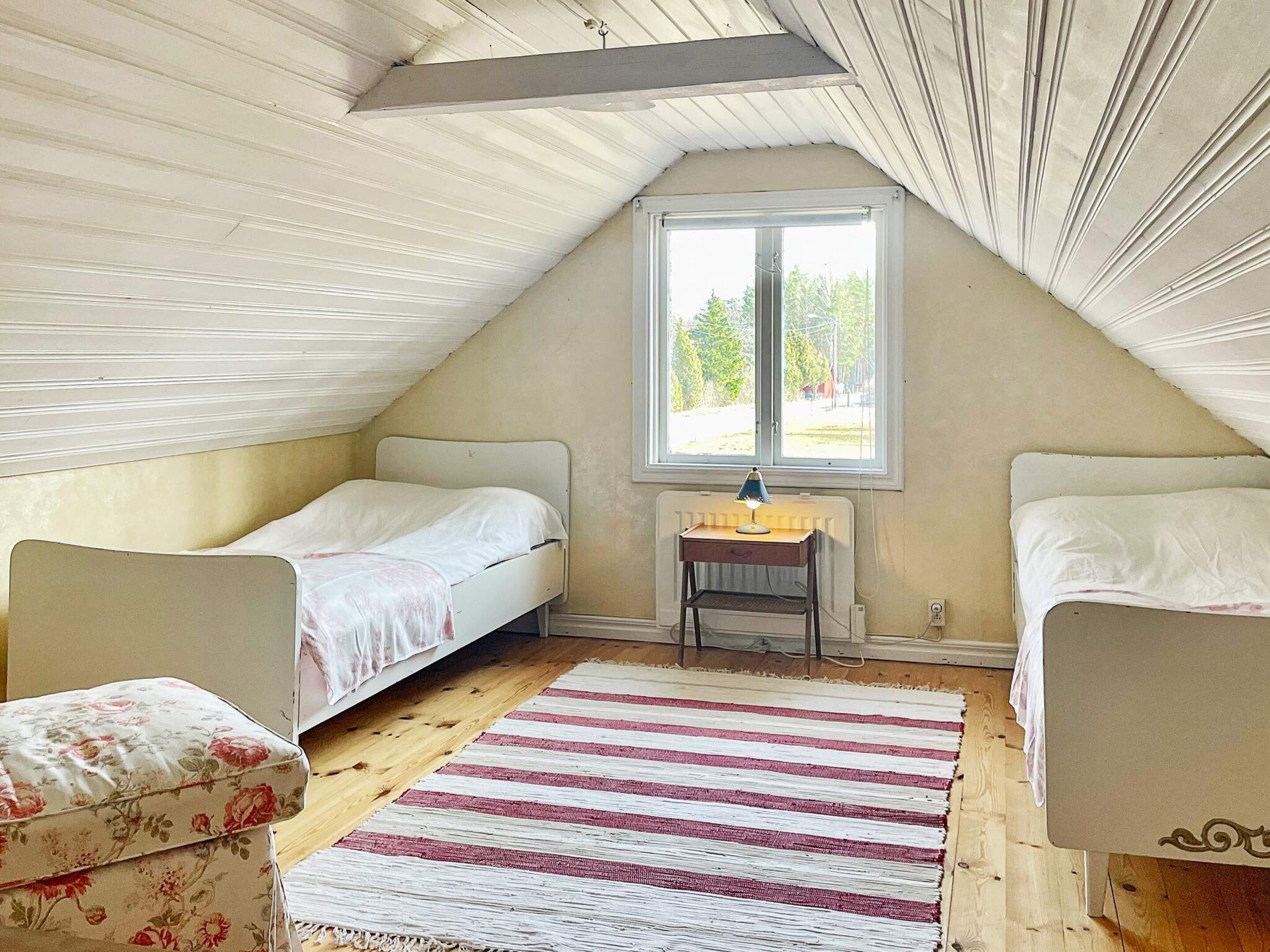 A small, cozy attic bedroom with two single beds, a wooden floor, a striped rug, and a window providing natural light.