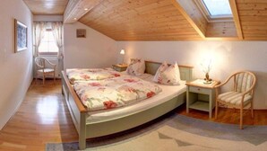 Ein Schlafzimmer mit Holzboden, einem Oberlicht, einem Bett mit Blumenmuster auf der Bettwäsche, einem Sessel und einem Nachttisch mit Lampe.