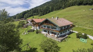 Eine zweistöckige Chalet mit geneigtem Dach, umgeben von üppigen grünen Feldern und einem bewaldeten Hang.