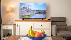A room with a television displaying a scenic view, a dining table with a bowl of fruit, a lamp, and a chair.