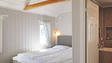 Quarto com cama, janela com cortinas, abajur e porta para o banheiro.